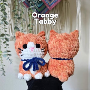 May include: Two crochet plush toys shaped like cats, labeled Orange Tabby. The front cat is orange and white with black eyes, a pink nose, and a dark blue ribbon. The back cat is solid orange with a dark blue collar. Both are made of soft yarn.