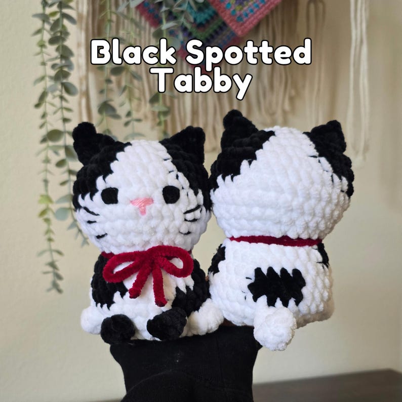 May include: Two plush crochet cat toys with black and white spotted patterns. The cat on the left features a pink stitched nose and a dark red yarn bow around its neck. The cat on the right shows its back with a dark red collar. Text reads Black Spotted Tabby.