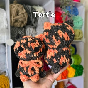 May include: Two handmade crochet plush toys featuring a mottled black and orange yarn pattern, resembling a tortoiseshell cat. The left plush has a small orange bow at the neck. The text "Tortie" is printed in white above the plush toys.