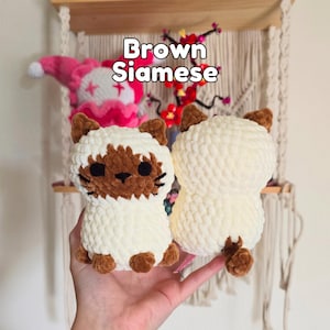 May include: Two crocheted plush cat toys made from soft, cream-colored yarn. The cat on the left features a brown face mask, brown ears, and brown paws. The cat on the right is cream-colored with brown ears and a small brown tail. Text reads Brown Siamese.