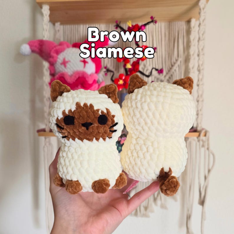 May include: Two crocheted plush cat toys made from soft, cream-colored yarn. The cat on the left features a brown face mask, brown ears, and brown paws. The cat on the right is cream-colored with brown ears and a small brown tail. Text reads Brown Siamese.