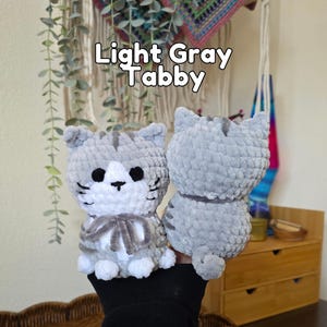 May include: Two light gray tabby cat plush toys made of soft yarn. One cat faces forward with a white chest, black eyes, and a gray bow, while the other shows its back. The text Light Gray Tabby is visible above the plush toys in white letters.