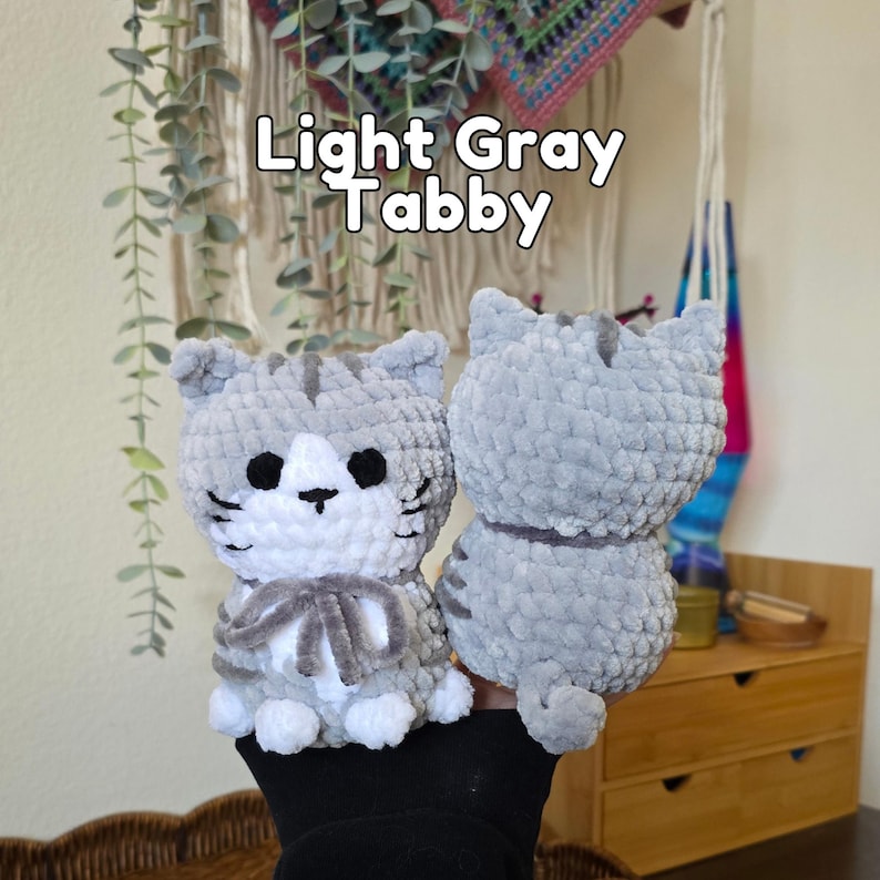 May include: Two light gray tabby cat plush toys made of soft yarn. One cat faces forward with a white chest, black eyes, and a gray bow, while the other shows its back. The text Light Gray Tabby is visible above the plush toys in white letters.