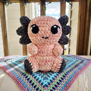 May include: A handmade crochet axolotl plush toy in shades of pink and gray, with large black eyes and a smiling expression. It sits on a colorful, patterned crochet blanket, showcasing a soft, textured design. The plush is a cute, whimsical creation.