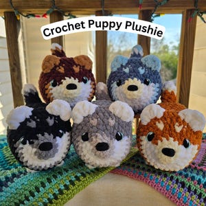 May include: Five handmade crochet puppy plushies in various colors: brown, blue, black, gray, and orange. Each plushie has white accents, black noses, and blue eyes. The text "Crochet Puppy Plushie" is above the plushies.