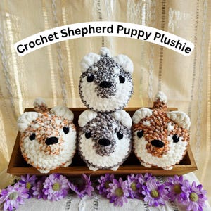 May include: Crochet Shepherd Puppy Plushies in a wooden tray. The plushies are white, brown, and gray with black eyes and noses. The text "Crochet Shepherd Puppy Plushie" is arched above the plushies. Purple flowers are at the bottom.