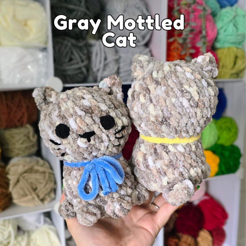 May include: Two crocheted cat figures made from gray, white, and brown mottled yarn. One cat features a blue yarn bow around its neck, while the other wears a yellow yarn collar. Both have black felt eyes and stitched noses. Text reads: Gray Mottled Cat.