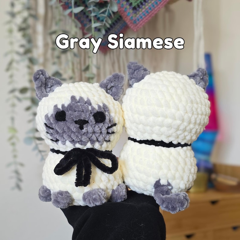 May include: Two crocheted plush cats with cream-colored bodies and gray accents on the ears, face, paws, and tail. The cat on the left features a black yarn bow around its neck and black embroidered eyes. The text Gray Siamese is visible above.