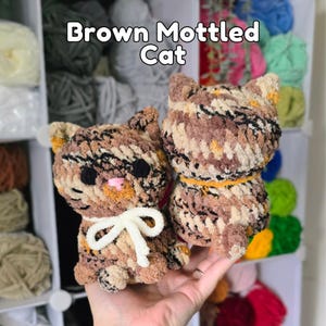 May include: Two crocheted cat figures made of brown, tan, and black mottled yarn. The left cat features a white yarn bow around its neck, while the right cat has an orange yarn collar. Both have black eyes and the text Brown Mottled Cat above.