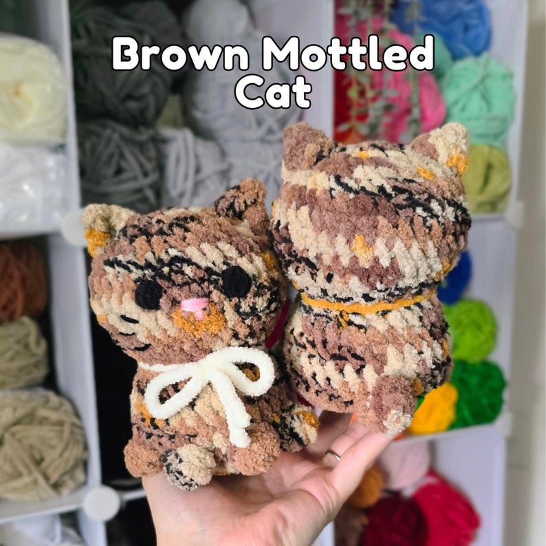 May include: Two crocheted cat figures made of brown, tan, and black mottled yarn. The left cat features a white yarn bow around its neck, while the right cat has an orange yarn collar. Both have black eyes and the text Brown Mottled Cat above.