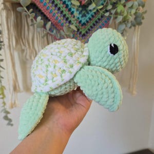 May include: A handmade plush turtle in soft green and white. The turtle's shell is white with a floral pattern, and its head, flippers, and body are a solid light green. It has a black eye and is made of a soft, textured material.