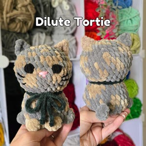 May include: A plush cat toy made of variegated gray and tan yarn with a soft texture. The cat features black circular eyes, a small pink stitched nose, and a dark green ribbon tied around its neck. The text Dilute Tortie is visible above.