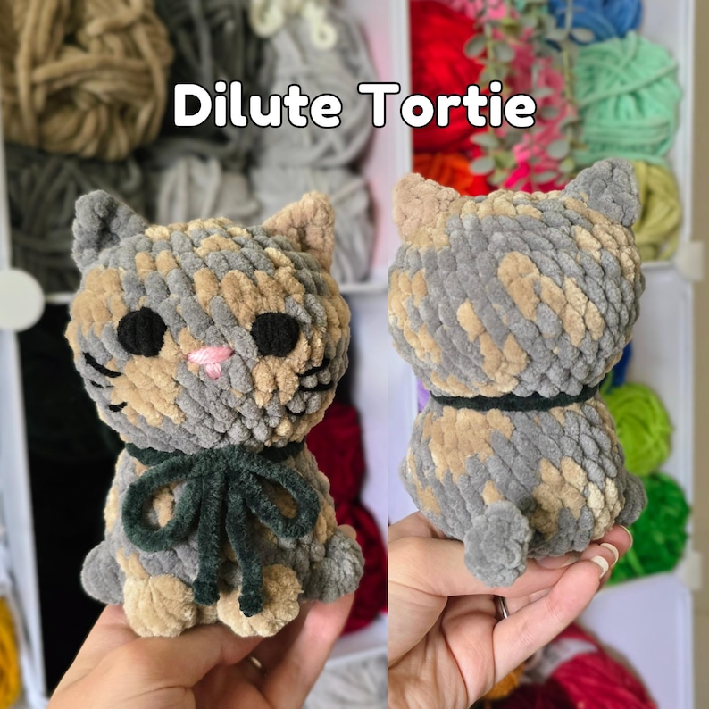 May include: A plush cat toy made of variegated gray and tan yarn with a soft texture. The cat features black circular eyes, a small pink stitched nose, and a dark green ribbon tied around its neck. The text Dilute Tortie is visible above.