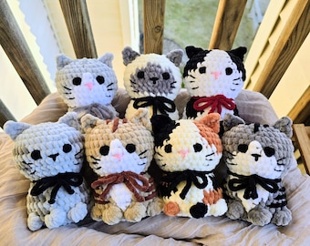 May include: A collection of seven small, handmade crochet cat plushies featuring soft, chunky yarn in various colors including gray, white, brown, black, and calico. Each cat has black safety eyes, a small stitched nose, and a tied ribbon.