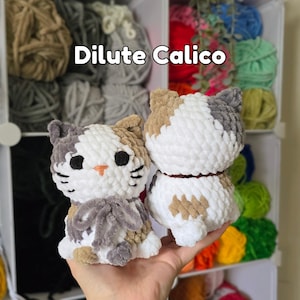 May include: Two plush cat figures made of soft, chunky white, grey, and tan yarn. The cat on the left faces forward with black eyes, a small orange nose, and a grey yarn bow. The cat on the right is shown from the back. Text reads: Dilute Calico.