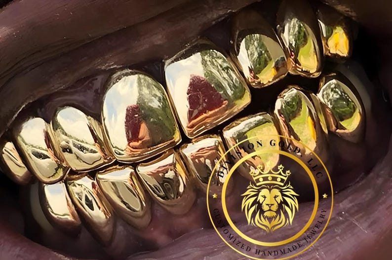 May include: Close-up of a set of gold teeth. The teeth are highly polished and reflect the surrounding environment. A circular logo with a lion's head and the text "BY JASON GOLD, LLC CUSTOMIZED HANDMADE JEWELRY" is visible.