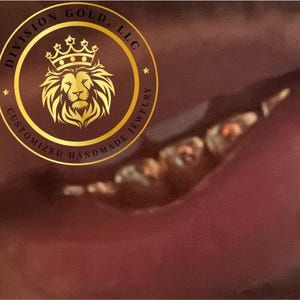 May include: A gold circular logo with a lion's head and crown, surrounded by the words "DIVISION GOLD, LLC" and "CUSTOMIZED HANDMADE JEWELRY". The background is a blurred, reddish-brown color.
