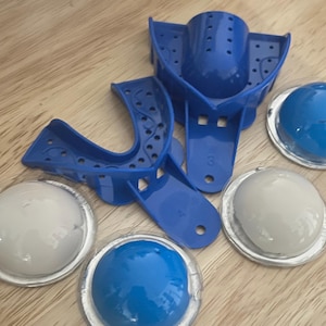 May include: A collection of dental impression trays and impression material. The trays are bright blue plastic, with perforated surfaces. The impression material is in clear, round containers, with blue and white contents. The items are arranged on a wooden surface.