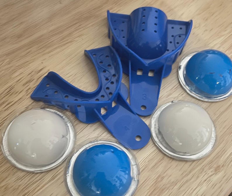 May include: A collection of dental impression trays and impression material. The trays are bright blue plastic, with perforated surfaces. The impression material is in clear, round containers, with blue and white contents. The items are arranged on a wooden surface.