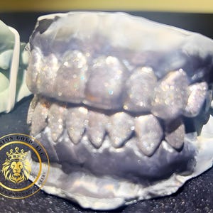 May include: A close-up of a dental mold with a set of teeth covered in glitter. The teeth are set in a gray mold, with a gold logo in the lower left corner. The logo reads "Division Gold, LLC, Customized Handmade Jewelry."