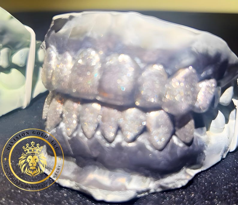 May include: A close-up of a dental mold with a set of teeth covered in glitter. The teeth are set in a gray mold, with a gold logo in the lower left corner. The logo reads "Division Gold, LLC, Customized Handmade Jewelry."