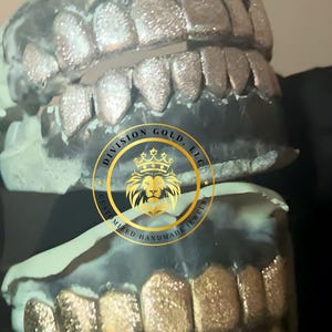 May include: Close-up of two sets of custom-made grillz. The top set is silver, and the bottom set is gold. The image includes the logo of Division Gold, LLC, which is a gold emblem with a lion's head and the text "Customized Handmade Jewelry."