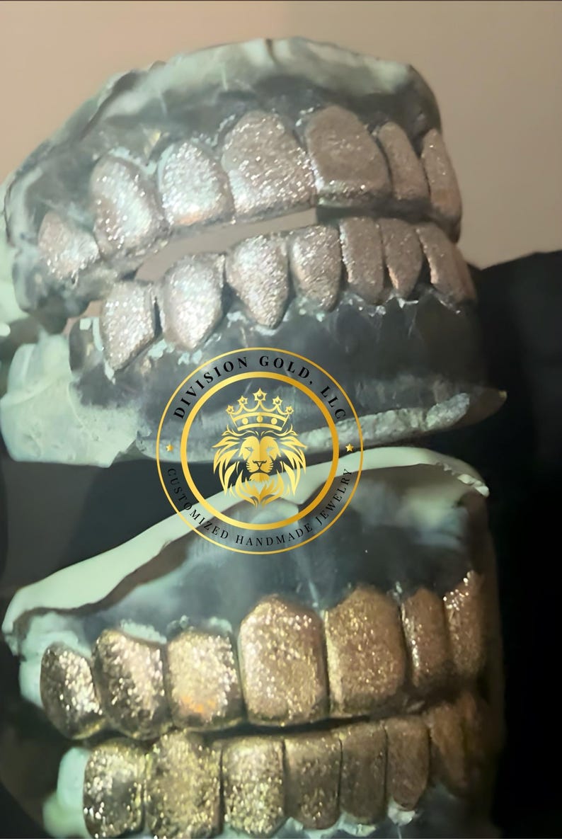 May include: Close-up of two sets of custom-made grillz. The top set is silver, and the bottom set is gold. The image includes the logo of Division Gold, LLC, which is a gold emblem with a lion's head and the text "Customized Handmade Jewelry."