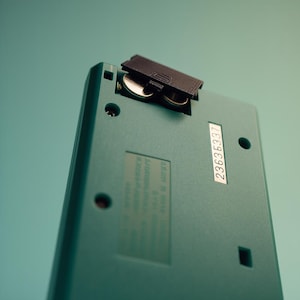May include: A close-up shot of the rear of a teal electronic device. The battery compartment is open, showing two silver batteries. A sticker with the number 23635337 is visible. The device has several small holes and a square indentation.