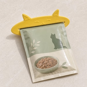 Può includere: Una clip per sacchetti in plastica giallo brillante a forma di testa di gatto con orecchie a punta, fissata su una busta rettangolare di cibo per gatti. La busta presenta uno sfondo verde salvia, una sagoma di gatto verde scuro e cibo umido.