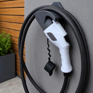 Op de afbeelding: Een witte en donkergrijze oplaadstekker voor elektrische voertuigen gemonteerd op een muurbeugel met een opgerolde zwarte oplaadkabel. De stekker heeft een zwarte beschermkap aan een golvend koord en de tekst MUSI|ART op de zijkant.