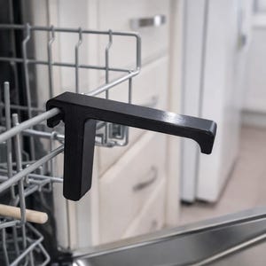 Può includere: Un regolatore nero per griglia lavastoviglie è attaccato alla griglia metallica all'interno di una lavastoviglie. Il regolatore ha una forma a T con un'impugnatura orizzontale e un supporto verticale. Lo sfondo mostra una cucina bianca.