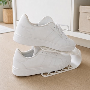 Pode incluir: Um par de ténis brancos empilhados verticalmente num organizador de sapatos de plástico branco. O organizador apresenta um design em camadas com uma base de padrão de grelha, permitindo que um sapato descanse sobre o outro para poupar espaço.