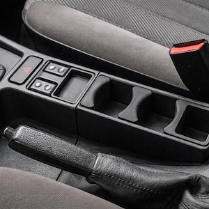 Puede incluir: Consola de coche negra con portavasos, interruptores y freno de mano. La consola es de plástico negro y está rodeada de asientos de tela gris. Se ve un botón rojo de luces de emergencia.