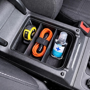 Op de afbeelding: Een auto-console-organizer met een gele rolmaat, een oranje kabel en handontsmettingsmiddel. De items zijn netjes gerangschikt in aparte compartimenten in de zwarte console, naast een autostoel met witte stiksels en een rode veiligheidsgordel.