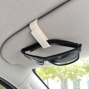 Könnte beinhalten: Ein cremefarbener Kunststoffclip an einer Sonnenblende im Fahrzeug, der eine Sonnenbrille mit schwarzem Rahmen hält. Der Clip hat ein U-förmiges Design, das über die Kante der Sonnenblende geschoben wird und eine sichere Halterung bietet.