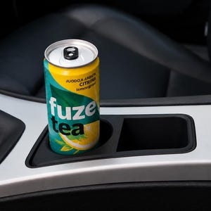 May include: A yellow and teal aluminium beverage can labelled Fuze Tea with the text JUODOJI ARBATA CITRINA lemongrass, featuring a graphic of a lemon slice, sitting inside a black plastic cup holder on a silver and black car centre console.