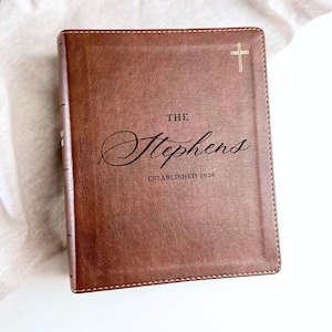 May include: Brown leather-bound book with the text "THE Stephens ESTABLISHED 2026" and a gold cross emblem. The book has a textured surface and is likely a journal or a personalized gift.