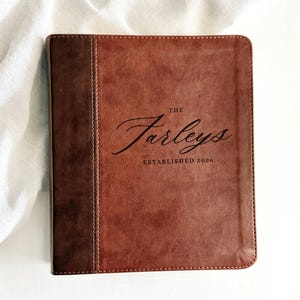 May include: A brown leather journal with the text "The Farleys ESTABLISHED 2026" in black script. The journal has a darker brown spine and is set against a white background with a white cloth.
