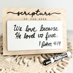 May include: Wooden sign with the words "scripture of the week" and a white dry-erase board. The board has the text "We love because He loved us first. I John 4:19" written on it. A black dry-erase marker is also present.