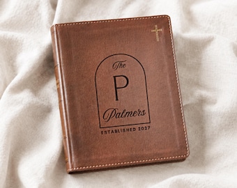 May include: A brown leather-like book cover featuring a gold cross in the top right corner. The center displays an arched design with the letter P, the text The Palmers, and the text ESTABLISHED 2027 printed in a dark, bold, sans-serif font.