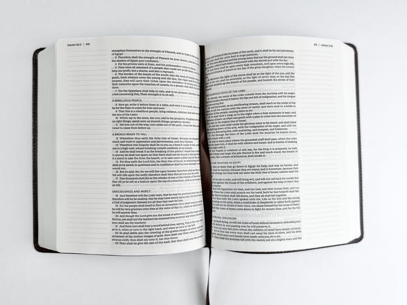 May include: An open book with white pages and black text, featuring a dark brown cover. The book is lying flat on a white surface. The text is small and densely packed, suggesting a religious or scholarly work.