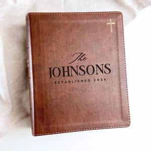 May include: A brown leather-bound book with the family name "The Johnsons" and "ESTABLISHED 2026" printed in black. A gold cross is in the upper right corner. The book is on a white surface.