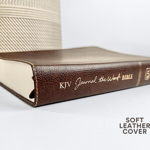 May include: A brown leather-bound Bible with the text "KJV Journal the Word BIBLE" in gold lettering. The book has a soft leather cover and cream-colored pages. A decorative beige woven vase is in the background.