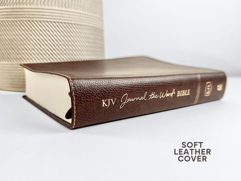 May include: A brown leather-bound Bible with the text "KJV Journal the Word BIBLE" in gold lettering. The book has a soft leather cover and cream-colored pages. A decorative beige woven vase is in the background.