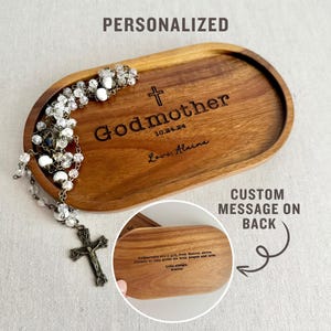 May include: Personalized wooden tray with the words "Godmother" and a cross engraved on the front. The back of the tray has a custom message. A white rosary with a cross pendant is on top of the tray.