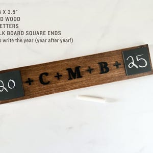 May include: A wooden sign with black lettering that reads "20 + C + M + B + 25". The sign has two small chalkboard squares on either end, one with the number "20" written in white chalk and the other with the number "25" written in white chalk. The sign is 45.1 cm by 8.9 cm.