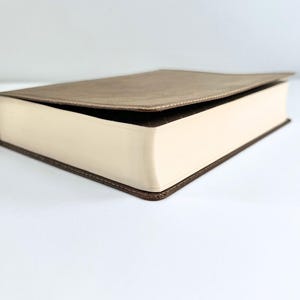 May include: A closed book with a light brown cover and cream-colored pages. The edges of the pages are visible, and the cover is slightly open. The book has a dark brown border.