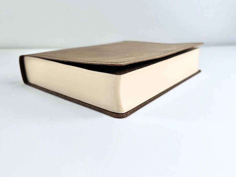 May include: A closed book with a light brown cover and cream-colored pages. The edges of the pages are visible, and the cover is slightly open. The book has a dark brown border.