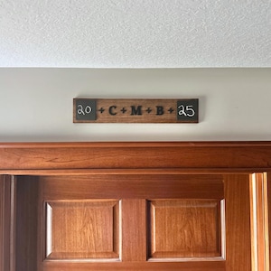 May include: A wooden sign with black lettering that reads "20 + C + M + B + 25" is mounted above a wooden door.