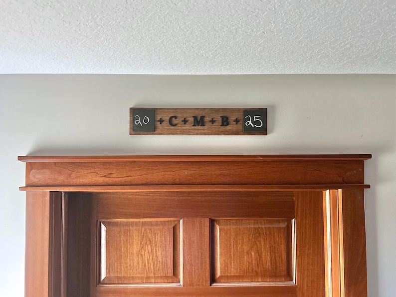 May include: A wooden sign with black lettering that reads "20 + C + M + B + 25" is mounted above a wooden door.